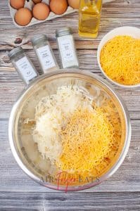 vertical photo of a mixing bowl with hash browns, cheese, spices and oil, with eggs, spices, oil and cheese behind it. All on a wooden surface