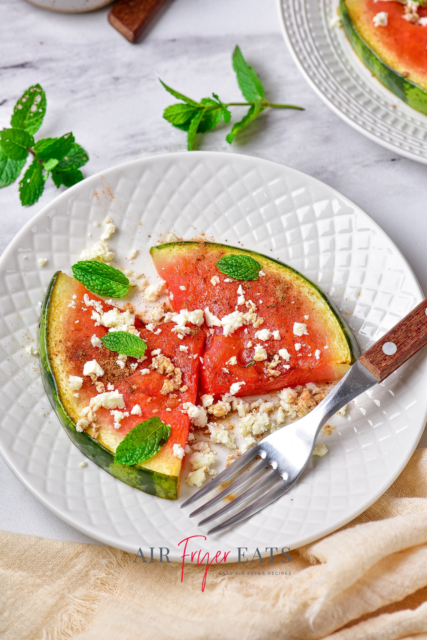 two pieces of air fried watermelon topped with fresh mint, dressing, and feta cheese, on a plate.