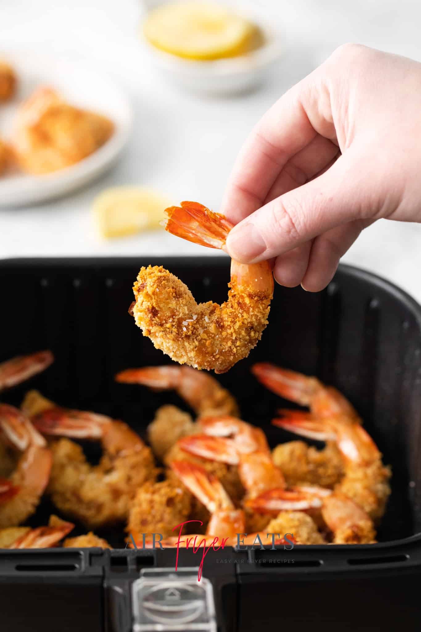 a hand holding up a breaded shrimp over an air fryer full of cooked shrimp.