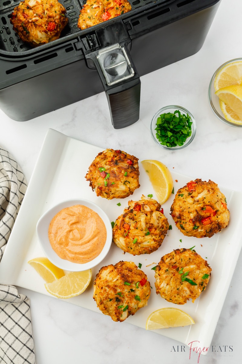 a rectangular plate of crab cakes with dipping sauce and lemons, next to an air fryer basket.