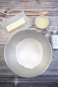 vertical photo of a mixing bowl with ingredients for the pizza base in it, with butter, olive oil and italian seasoning around the bowl. All on a wooden surface