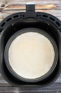 vertical photo of a pie pan in an air fryer basket with the pizza base cooked for 5 minutes