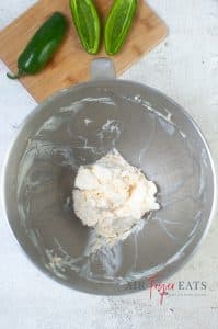 vertical photo of a large mixing bowl with whipped garlic powder, cream cheese and shredded cheese whipped. A wooden cutting board with jalapenos on it is in the background