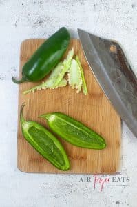 vertical photo of a wooden cutting board with 2 jalapenos, one intact and one sliced in half with the inside cleaned.