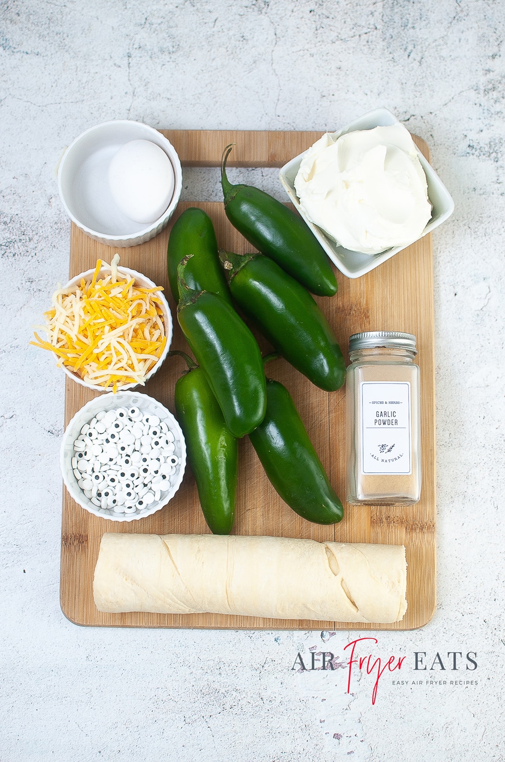 vertical photo of a wooden board with the ingredients for Air Fryer Mummy Poppers - jalapenos, crescent rolls, cream cheese, shredded cheese, garlic powder, egg, candy eyeballs