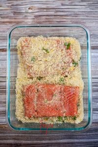vertical photo of a glass dish with the breadcrumb mixture and 2 pieces of salmon being coated. On a wooden surface