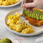 a plate of air fryer scrambled eggs with avocado toast. a fork is picking up a piece of egg.