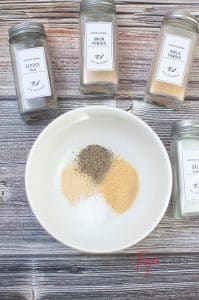 vertical photo of the spices for air fryer bacon wrapped chicken salt, pepper, onion powder, and garlic powder. On a wooden surface