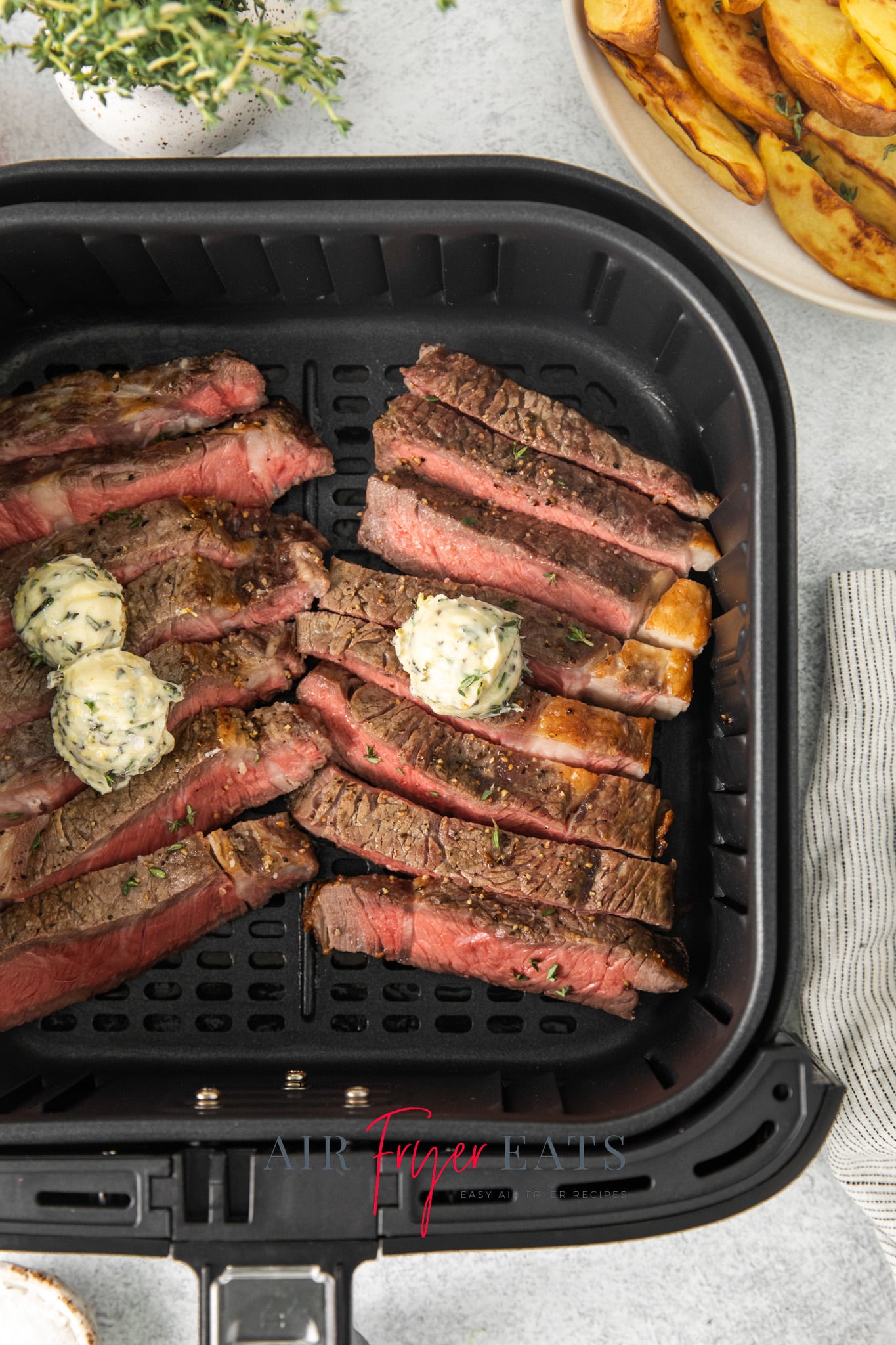 sliced ribeye steaks in an air fryer basket with herb butter on top.