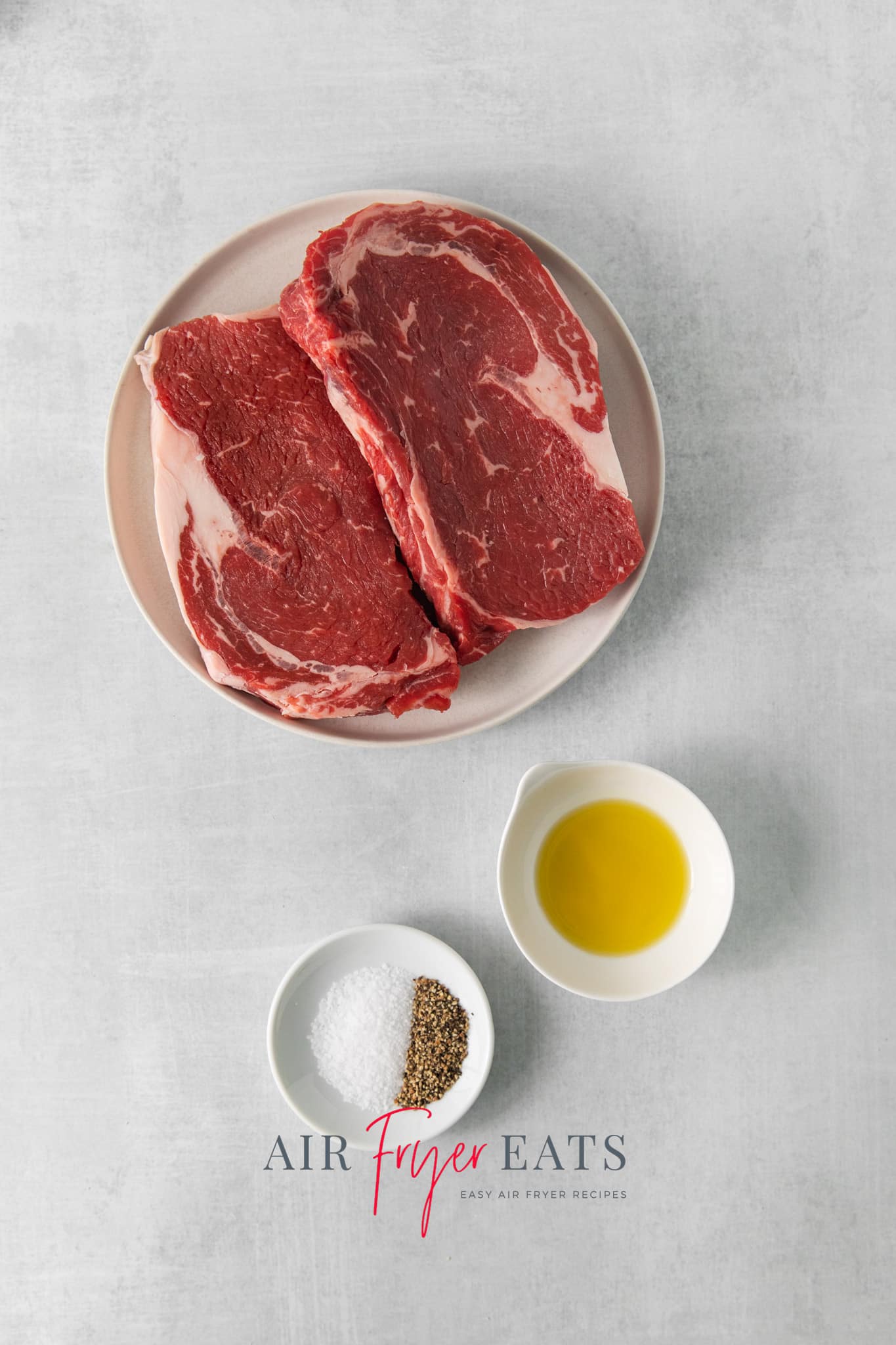 two ribeye steaks on a plate next a bowl of olive oil and another bowl of salt and pepper.