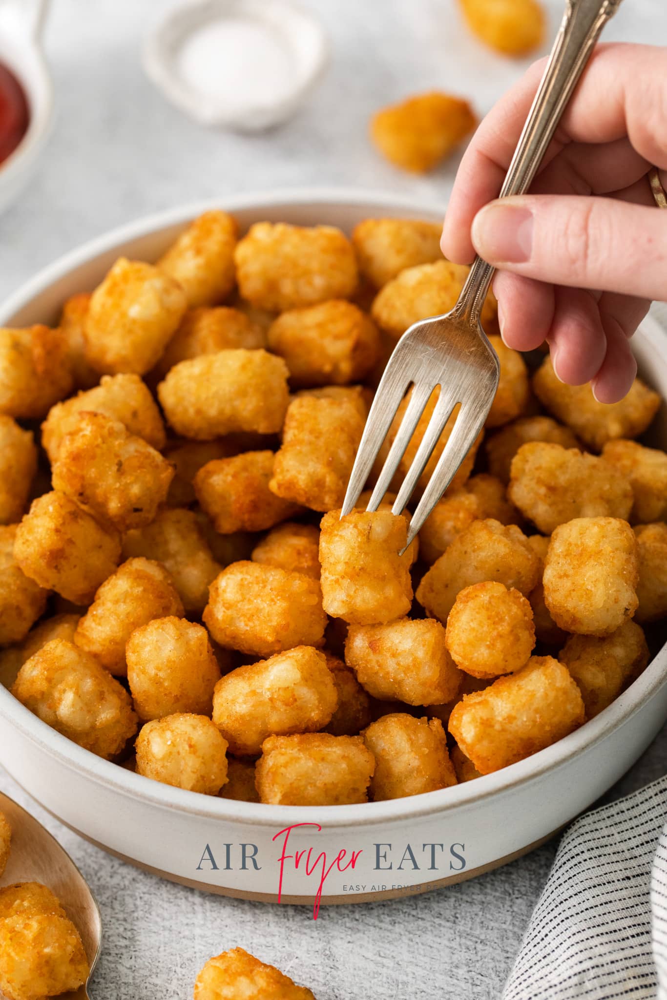 a bowl of crispy air fryer tater tots. A hand is picking one up with a fork.