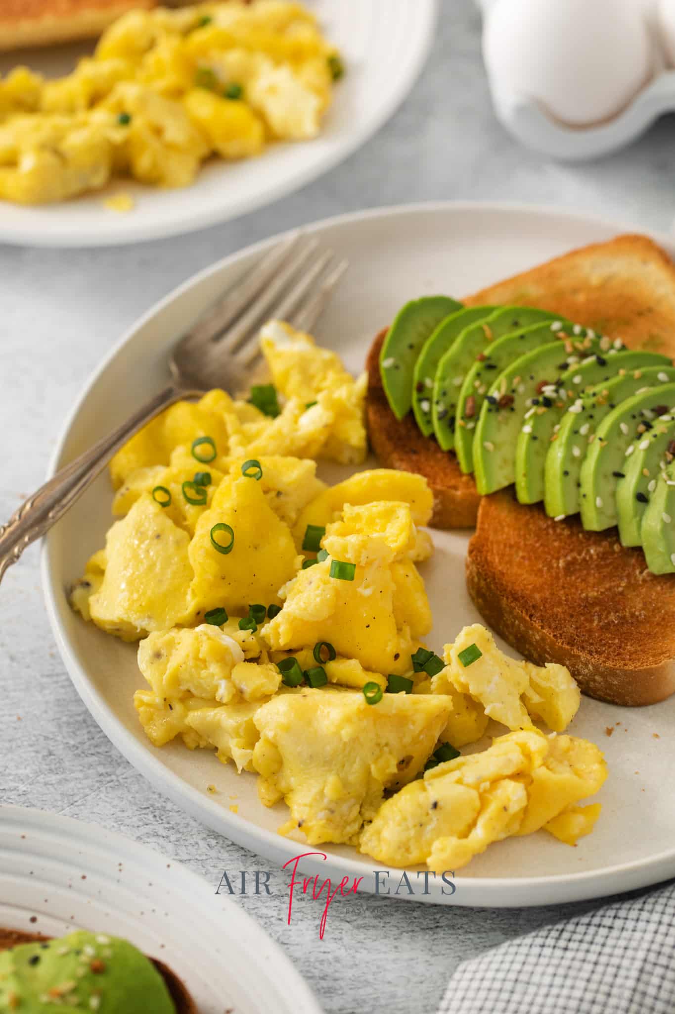 a plate of scrambled eggs that were cooked in an air fryer, garnished with green onion and served with a slice of toast with avocado.