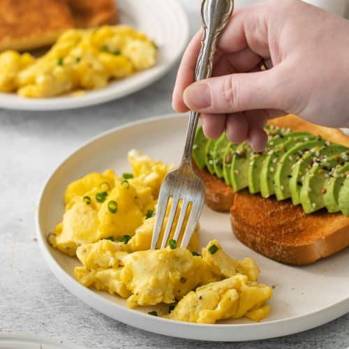 a hand holding a fork, enjoying a breakfast plate of avocado toast and air fryer scrambled eggs.