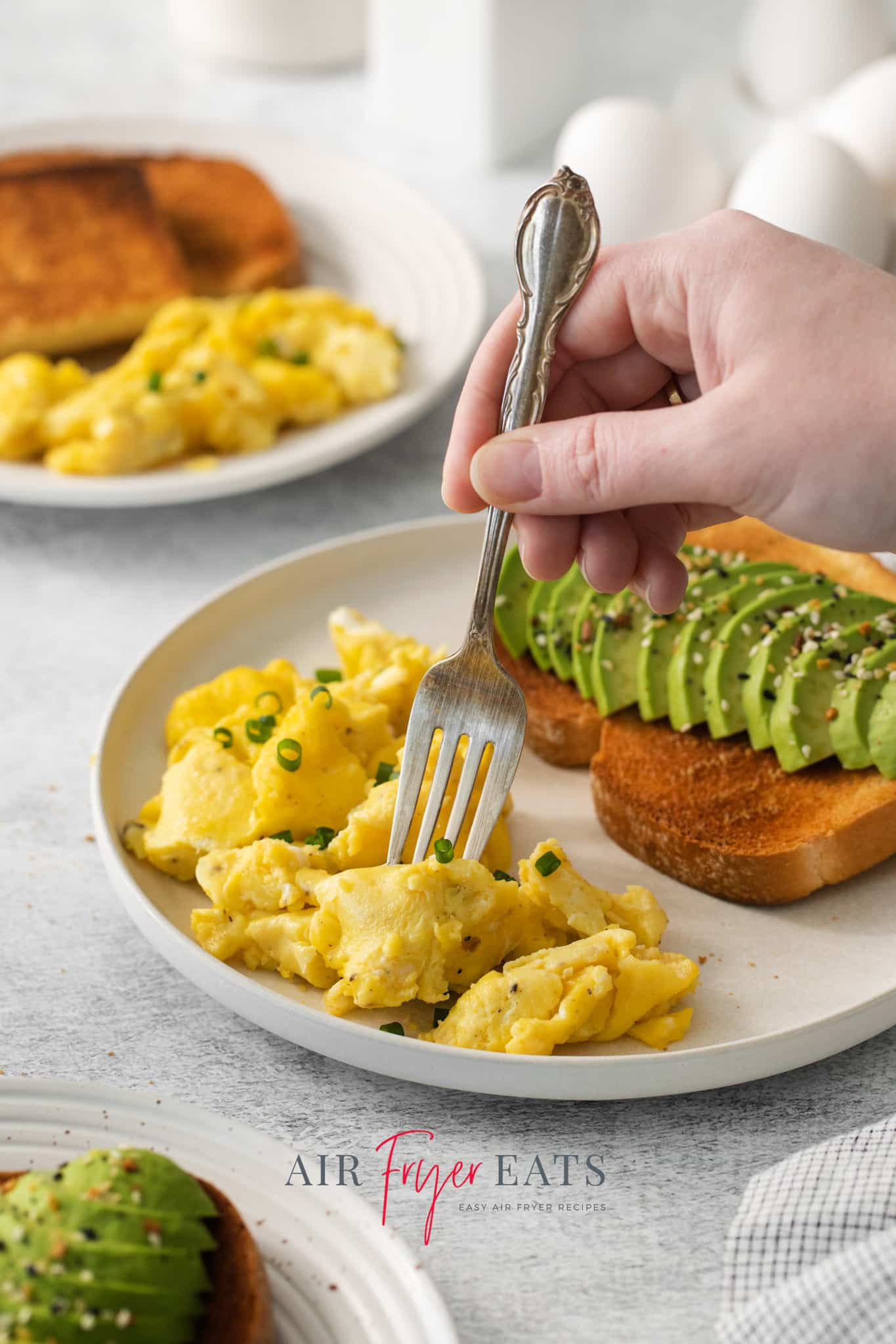 a hand holding a fork, enjoying a breakfast plate of avocado toast and air fryer scrambled eggs.