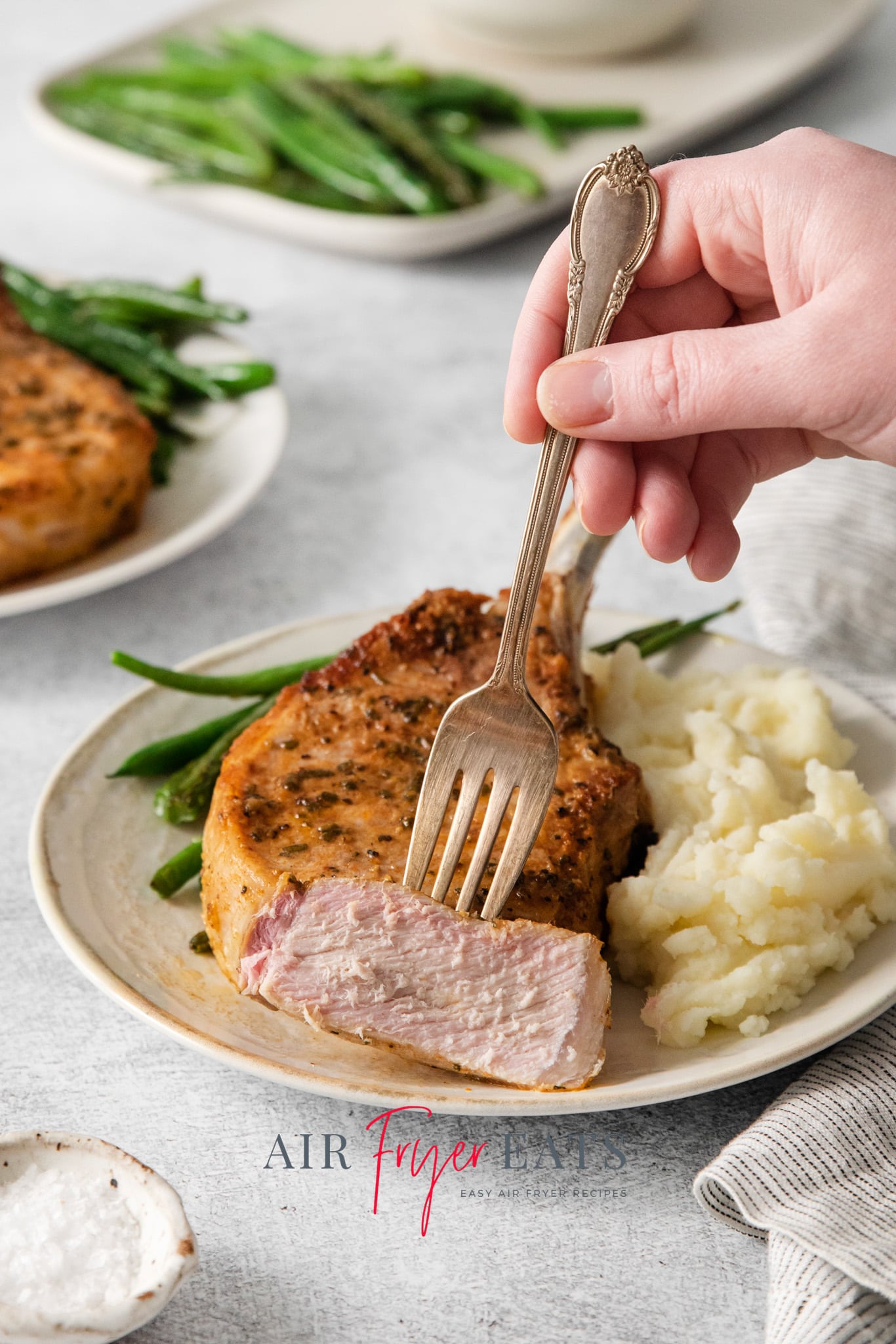 a dinner plate of pork chop with potatoes and green beans. A slice is held by a fork to show the juicy center.