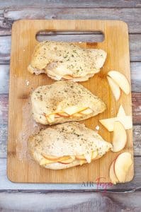 vertical photo of three chicken breasts on a wooden cutting board sliced and stuffed with cheese and apple slices