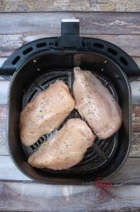 vertical photo of three seasoned chicken breasts in the air fryer basket ready to cook