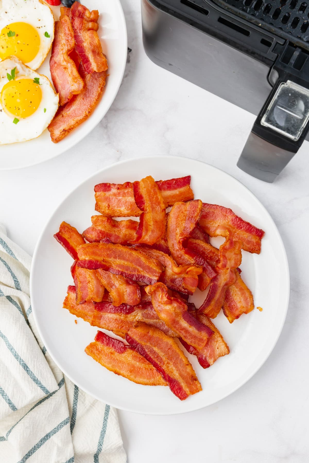 A plate full of crispy air fryer bacon is alongside a plate of fried eggs and bacon and a large, black air fryer.