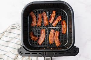 Sliced of bacon are cooked and crispy, they lay in the bottom of an air fryer basket.