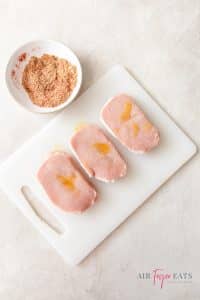 A plastic cutting board has three raw pork chops laid atop it. There is a small white bowl of spices nearby.