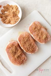 Three large raw pork chops are atop a plastic cutting board and covered with a spice mix.