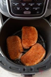 Three raw seasoned pork chops are placed in a round air fryer basket.