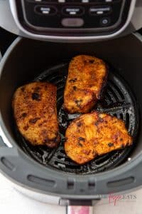 Three cooked air fryer pork chops are in an air fryer basket. They are crispy and a rich red orange color.