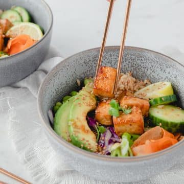 A close up picture of an air fryer poke bowl. Someone is reaching into the bowl with a pair of chopsticks and grabbing a piece of tofu.