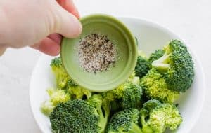A small green bowl of salt and pepper is sprinkled over a bowl of washed and cut broccoli florets.