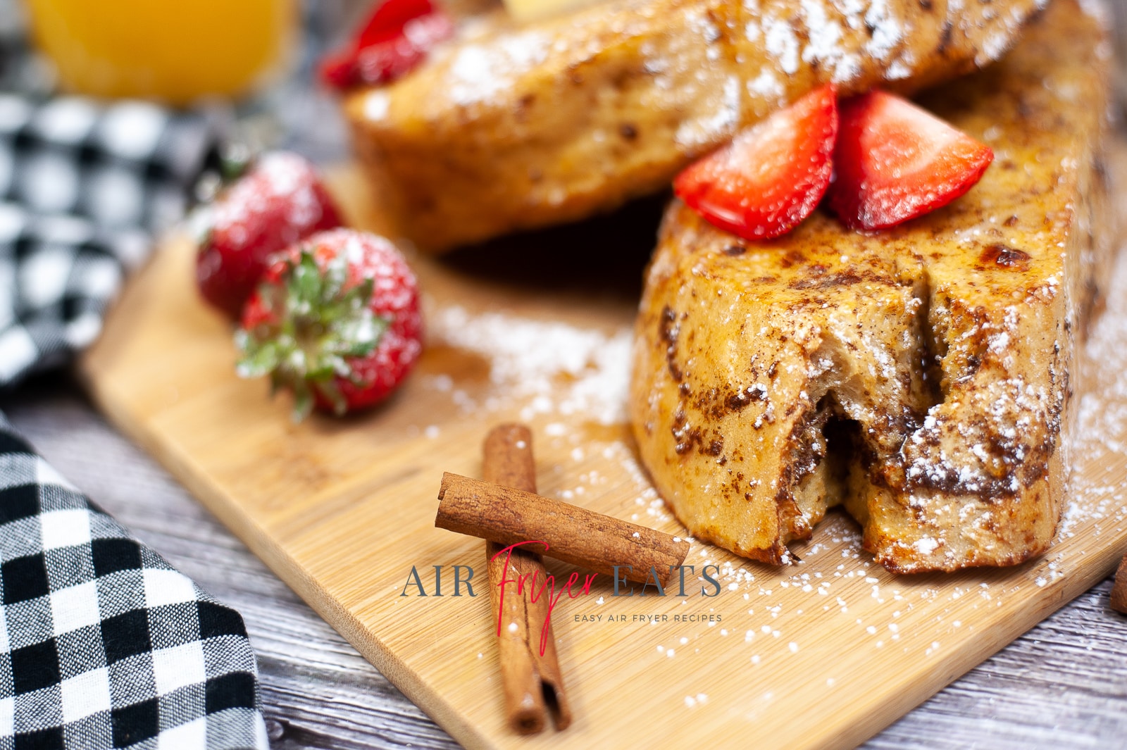 A close-up shot of two thick slices of air fryer french toast. They are covered in cinnamon, sliced strawberries, powdered sugar, and maple syrup.