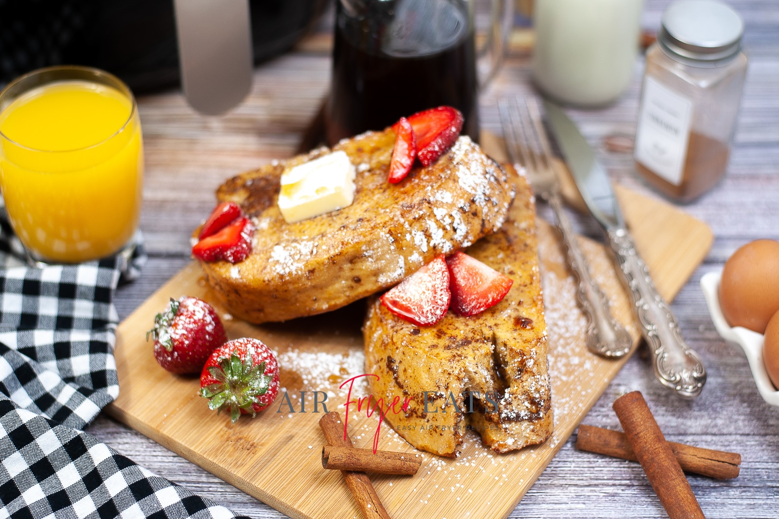 Two slices of air fryer french toast are covered with fresh strawberries, a slice of butter, and powdered sugar. They are placed on a wooden cutting board and there is a large container of maple syrup and a glass of orange juice nearby.