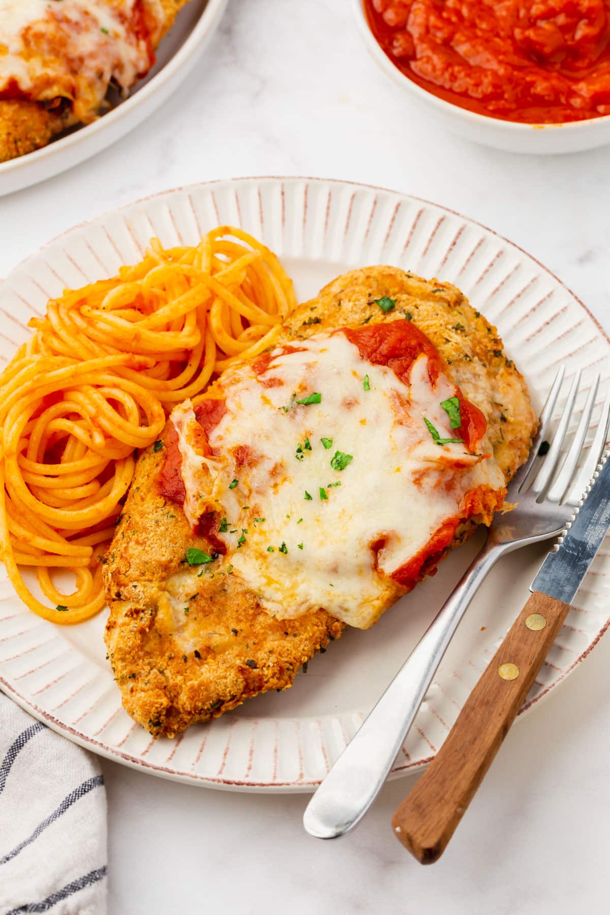 A plate is filled with spaghetti with a red sauce and a breaded air fryer chicken parmesan cutlet topped with marinara sauce, mozarella cheese, and freshly chopped parsley. A silver fork and knife lie on the right side of the white plate.