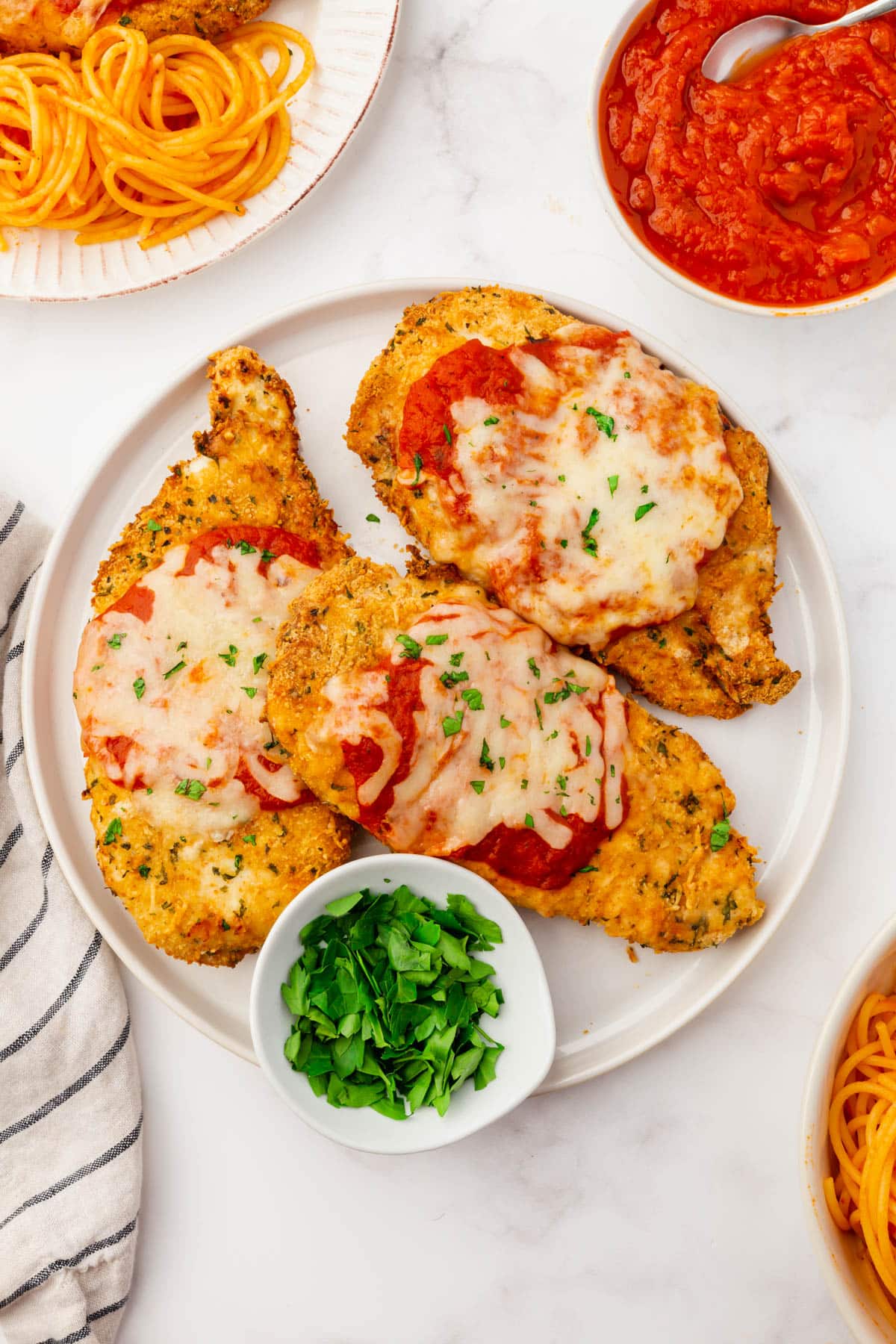 Three air fryer chicken parmesan cutlets lie on a white plate. Marinara sauce, freshly chopped parsley, and spaghetti with a red sauce surround the plate.