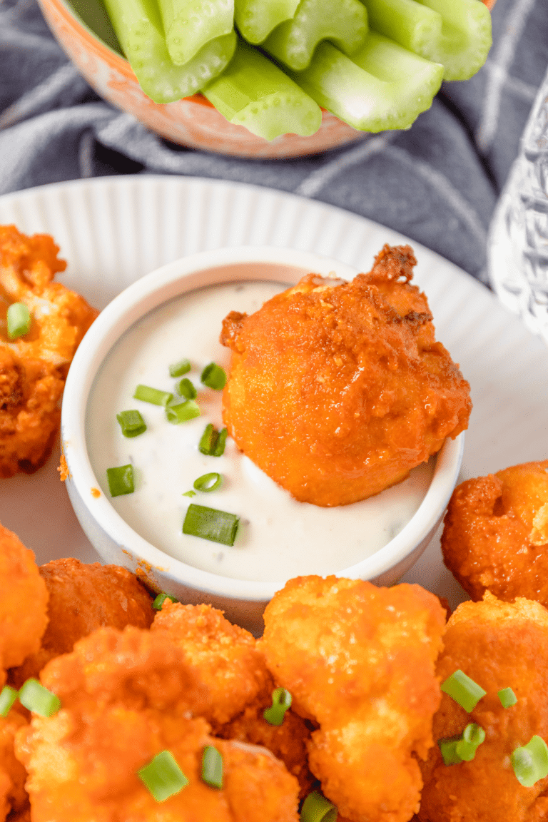 A red-orange buffalo cauliflower floret dipped in a white ranch sauce. A plate is filled with air fryer buffalo cauliflower and green celery sticks are piled in a bowl nearby.