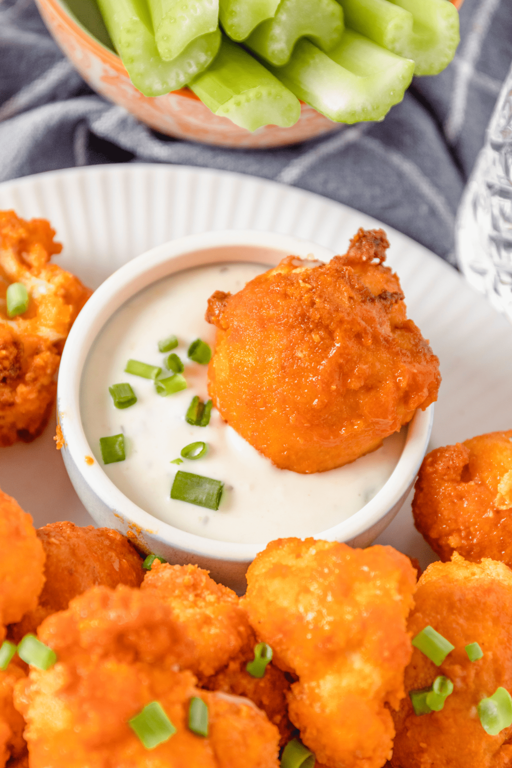 A red-orange buffalo cauliflower floret dipped in a white ranch sauce. A plate is filled with air fryer buffalo cauliflower and green celery sticks are piled in a bowl nearby.