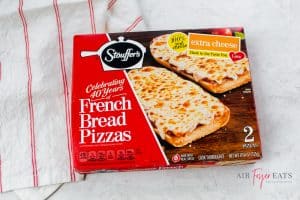 A red box of frozen Stouffer’s French bread pizza with extra cheese sits on a kitchen countertop next to a red and white striped kitchen towel.