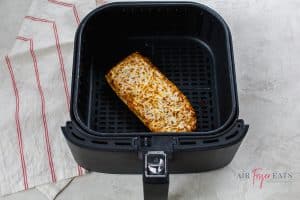One piece of cooked Stouffer’s French bread pizza lies in a black air fryer basket. The cheese pizza is golden-brown and crispy. The air fryer lies on a white and red striped kitchen towel.