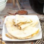 Air fryer chicken quesadillas are plated on a white, rectangular plate. A black air fryer can be seen in the background, along with a small bowl of red salsa.