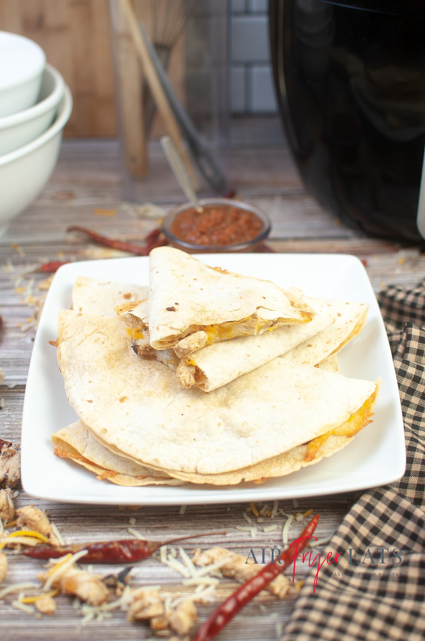 Air fryer chicken quesadillas are plated on a white, rectangular plate. A black air fryer can be seen in the background, along with a small bowl of red salsa.