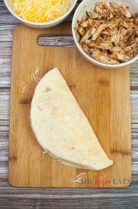 A large flour tortilla is folded in half and placed on a wooded cutting board on a kitchen counter. There is a bowl filled with yellow and white shredded cheese and a bowl filled with chicken peices nearby.
