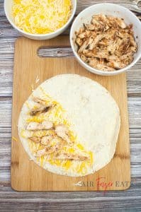 Half of a large flour tortilla is covered with cheese and chicken and placed on a wooded cutting board on a kitchen counter. There is a bowl filled with yellow and white shredded cheese and a bowl filled with chicken peices nearby.