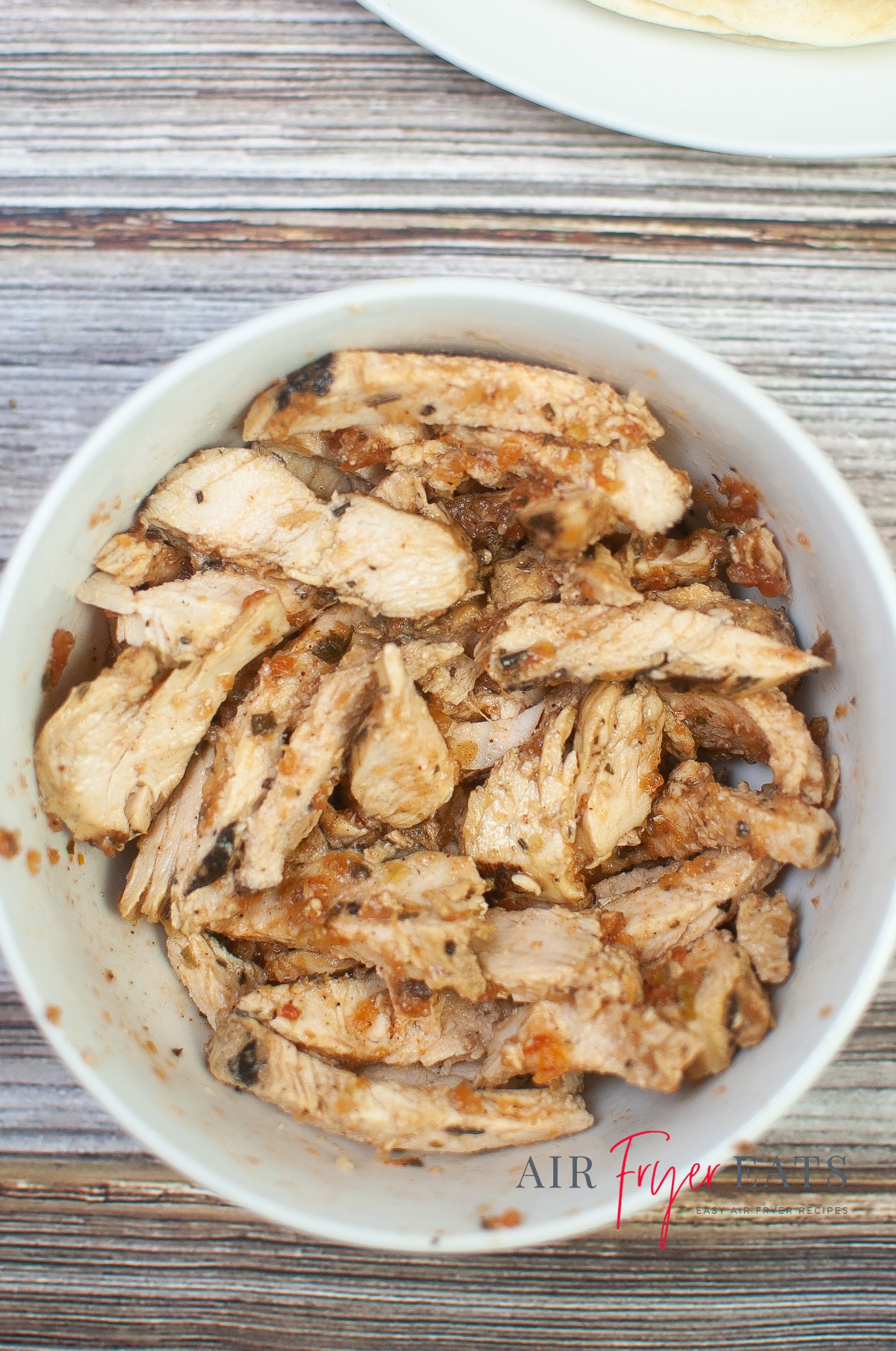 A close-up shot of a large white bowl filled with cooked chicken pieces and flakes of red salsa. A plate full of flour tortillas can be seen in the background.