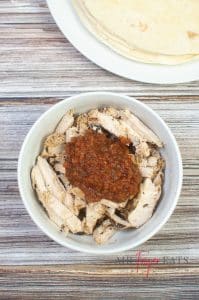 A large white bowl is filled with peices of cooked chicken. A large spoonful of red salsa is atop the chicken. A plate full of large flour tortillas is in the background.
