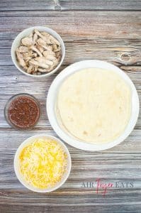 Atop a wooden table there are bowls of ingredients: a bowl of cooked chicken peices, a bowl of red salsa, and a bowl of shredded yellow and white cheese. A large plate full of flour tortillas is nearby.