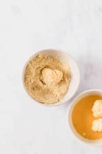 A small cauliflower floret is covered with flour inside a small bowl filled with flour. Nearby, another small bowl is filled with a butter and buffalo sauce mixture.
