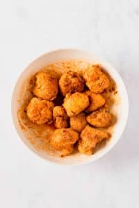 A large bowl is filled with raw cauliflower florets covered in buffalo sauce. The bowl is placed on a clean, white kitchen table.