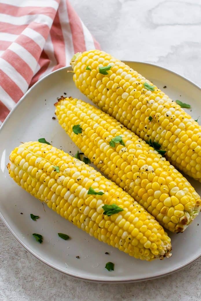 Three ears of air fryer corn on the cob are sprinkled with salt and pepper and parsley. The yellow corn is tender and juicy and sits on a white plate.