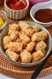 Golden-brown homemade air fryer tater tots are piled in a ceramic bowl. Dipping sauces like ketchup, barbeque sauce, and mustard are in the background.