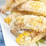 Golden-yellow Mexican street corn made in the air fryer is covered with white Parmesan cheese, red spices, and green flakes. The corn pieces are piled on top of each other on a white plate.