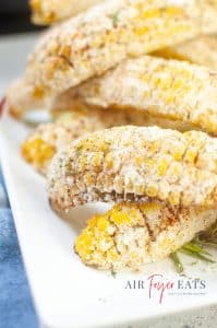 Golden-yellow Mexican street corn made in the air fryer is covered with white Parmesan cheese, red spices, and green flakes. The corn pieces are piled on top of each other on a white plate.